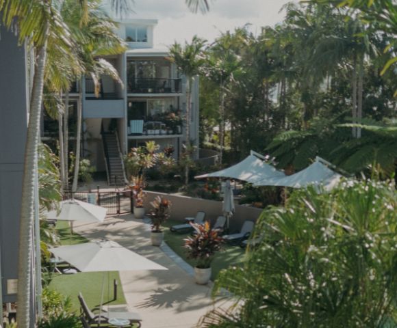 Lush tropical courtyard with palm trees and sunlit pathways. Lounge chairs and umbrellas create a serene, resort-like atmosphere beside modern buildings.