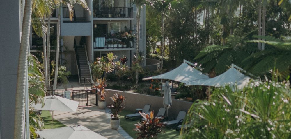 Lush tropical courtyard with palm trees and sunlit pathways. Lounge chairs and umbrellas create a serene, resort-like atmosphere beside modern buildings.