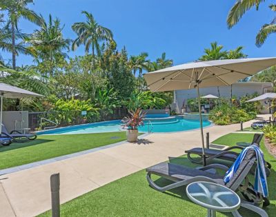 Sunny resort pool area with loungers, umbrellas, and lush tropical plants. Bright blue sky adds to the relaxing, vacation atmosphere.