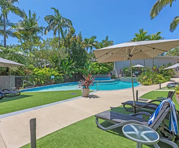 Sunny resort pool area with loungers, umbrellas, and lush tropical plants. Bright blue sky adds to the relaxing, vacation atmosphere.
