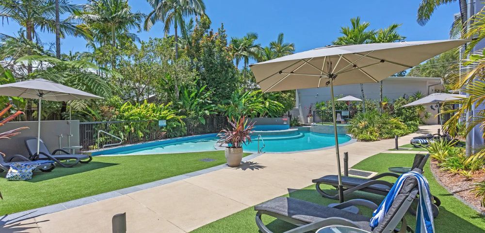 Sunny resort pool area with loungers, umbrellas, and lush tropical plants. Bright blue sky adds to the relaxing, vacation atmosphere.