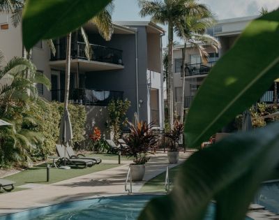 Tropical resort with palm trees and a pool surrounded by modern gray buildings. Sun loungers and lush greenery create a relaxing atmosphere.