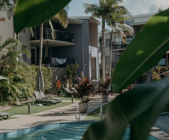 Tropical resort with palm trees and a pool surrounded by modern gray buildings. Sun loungers and lush greenery create a relaxing atmosphere.