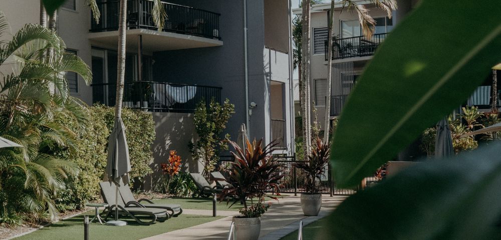 Tropical resort with palm trees and a pool surrounded by modern gray buildings. Sun loungers and lush greenery create a relaxing atmosphere.
