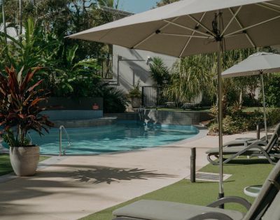 A serene outdoor pool area with lounge chairs and umbrellas, surrounded by lush green plants and trees. Sunlight casts soft shadows, creating a relaxing atmosphere.
