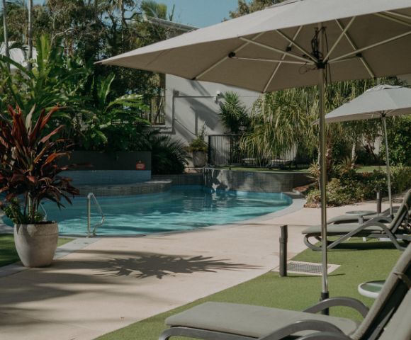 A serene outdoor pool area with lounge chairs and umbrellas, surrounded by lush green plants and trees. Sunlight casts soft shadows, creating a relaxing atmosphere.