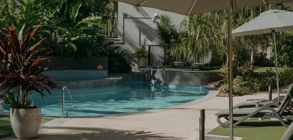 A serene outdoor pool area with lounge chairs and umbrellas, surrounded by lush green plants and trees. Sunlight casts soft shadows, creating a relaxing atmosphere.