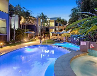 Luxurious pool area at dusk with illuminated water, surrounded by palm trees and modern buildings. Lounge chairs and umbrellas create a tranquil, inviting atmosphere.