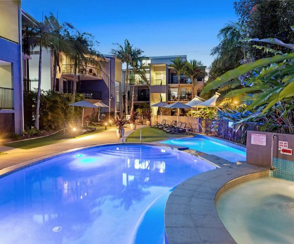 Luxurious pool area at dusk with illuminated water, surrounded by palm trees and modern buildings. Lounge chairs and umbrellas create a tranquil, inviting atmosphere.