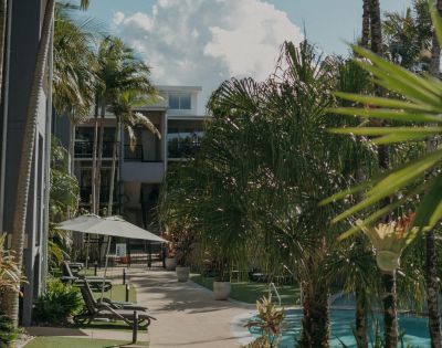 A tropical courtyard with lush palm trees surrounds a tranquil pool. Sun loungers and a parasol offer a relaxing atmosphere under a bright, cloudy sky.