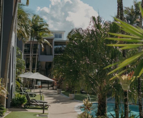 A tropical courtyard with lush palm trees surrounds a tranquil pool. Sun loungers and a parasol offer a relaxing atmosphere under a bright, cloudy sky.