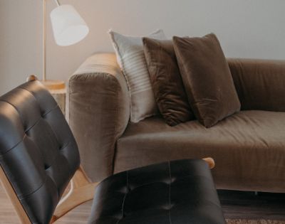A cozy living room features a brown velvet sofa with plush cushions, a modern black leather chair, and a soft-lit floor lamp, offering a warm and inviting ambiance.