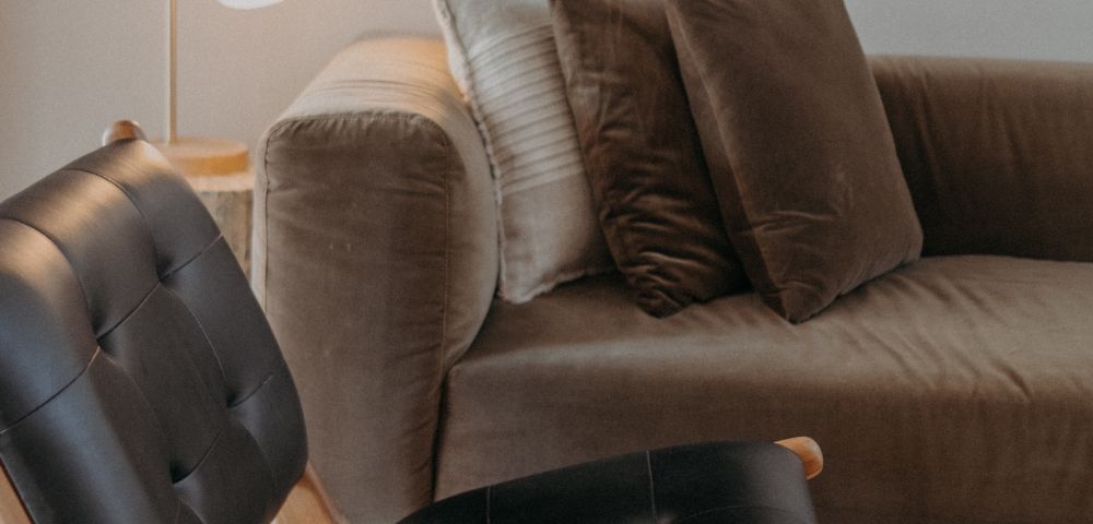 A cozy living room features a brown velvet sofa with plush cushions, a modern black leather chair, and a soft-lit floor lamp, offering a warm and inviting ambiance.