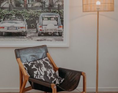 Contemporary living room with a leather chair, patterned cushion, and tall lamp. Background features a framed photo of vintage cars amidst greenery. Cozy ambiance.