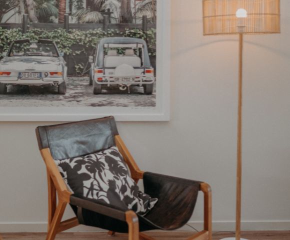 Contemporary living room with a leather chair, patterned cushion, and tall lamp. Background features a framed photo of vintage cars amidst greenery. Cozy ambiance.