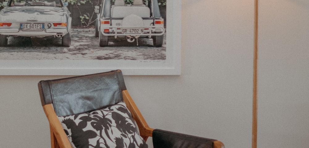 Contemporary living room with a leather chair, patterned cushion, and tall lamp. Background features a framed photo of vintage cars amidst greenery. Cozy ambiance.