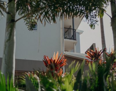 A white building with a small balcony is partially obscured by tropical plants. Palm trees and red-leaved shrubs create a lush, serene atmosphere.