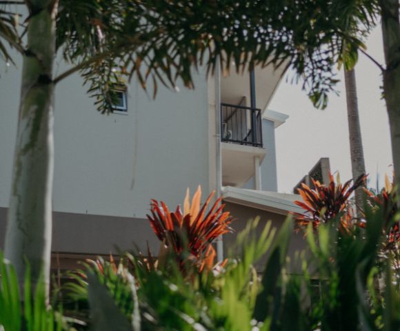 A white building with a small balcony is partially obscured by tropical plants. Palm trees and red-leaved shrubs create a lush, serene atmosphere.