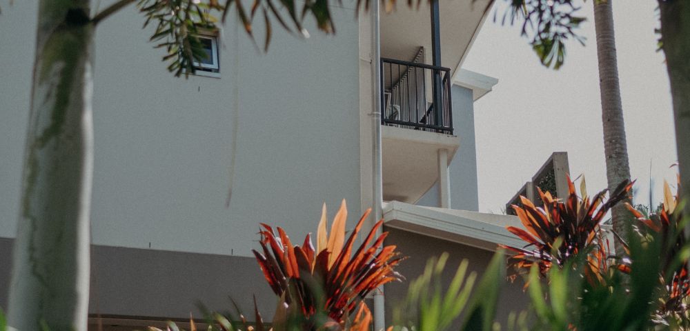 A white building with a small balcony is partially obscured by tropical plants. Palm trees and red-leaved shrubs create a lush, serene atmosphere.