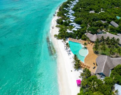 Aerial view of a tropical beach with turquoise waters, white sand, palm trees, luxury villas, and a pool. Lush greenery surrounds the tranquil setting.