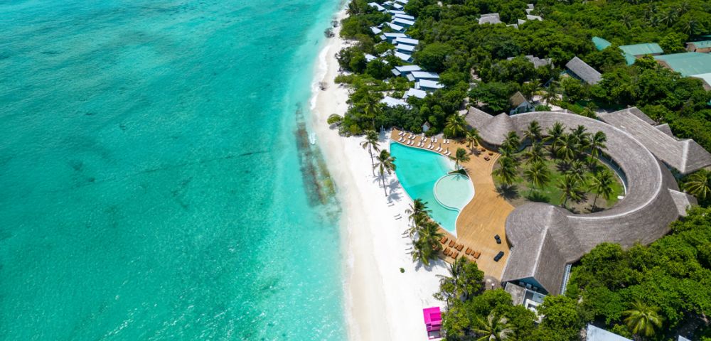 Aerial view of a tropical beach with turquoise waters, white sand, palm trees, luxury villas, and a pool. Lush greenery surrounds the tranquil setting.