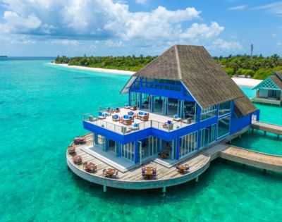 A blue, thatched-roofed overwater restaurant sits above clear turquoise ocean water. It's surrounded by tropical greenery under a partly cloudy sky, evoking luxury.