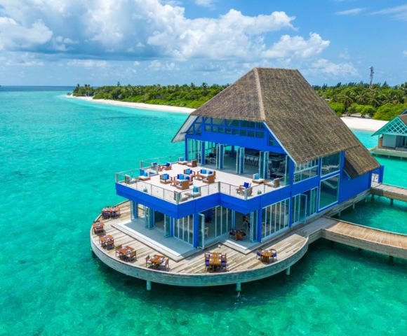 A blue, thatched-roofed overwater restaurant sits above clear turquoise ocean water. It's surrounded by tropical greenery under a partly cloudy sky, evoking luxury.