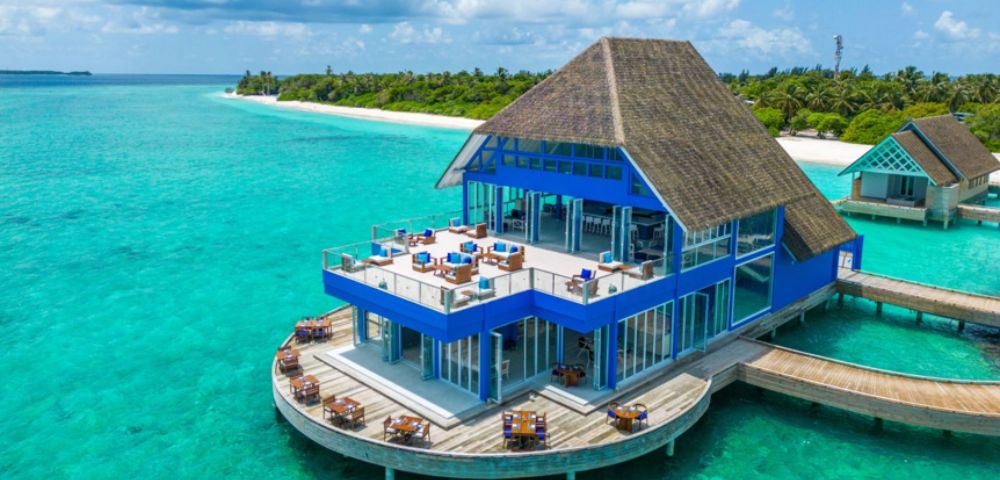 A blue, thatched-roofed overwater restaurant sits above clear turquoise ocean water. It's surrounded by tropical greenery under a partly cloudy sky, evoking luxury.