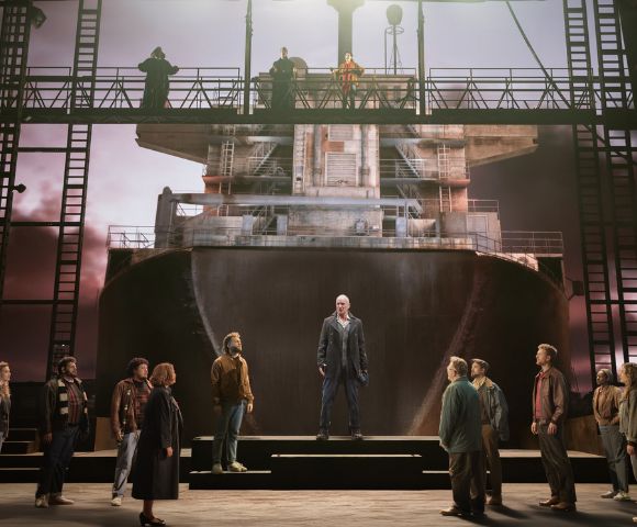 A group of people stands on a stage set resembling a ship's hull under a dramatic sky. A person stands centered and elevated, adding a solemn tone.