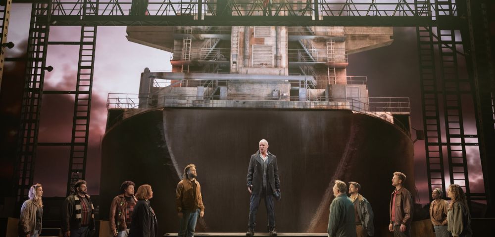 A group of people stands on a stage set resembling a ship's hull under a dramatic sky. A person stands centered and elevated, adding a solemn tone.