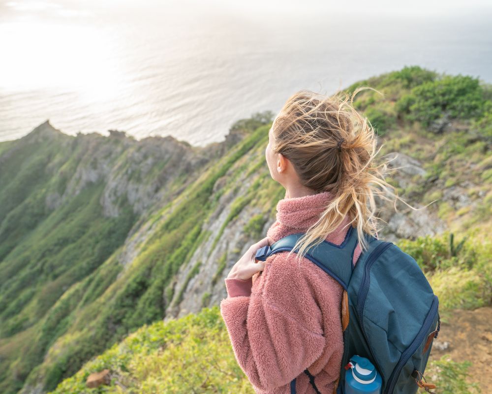 A woman with a backpack gazes at a scenic coastal view from a grassy cliff. Her windswept hair and peaceful expression convey a sense of adventure.