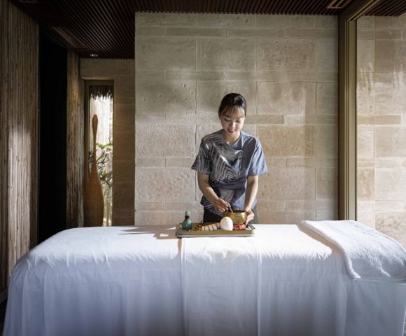A serene room with a massage table draped in white linens. A person arranges spa items with care, surrounded by soft natural light and wooden decor.