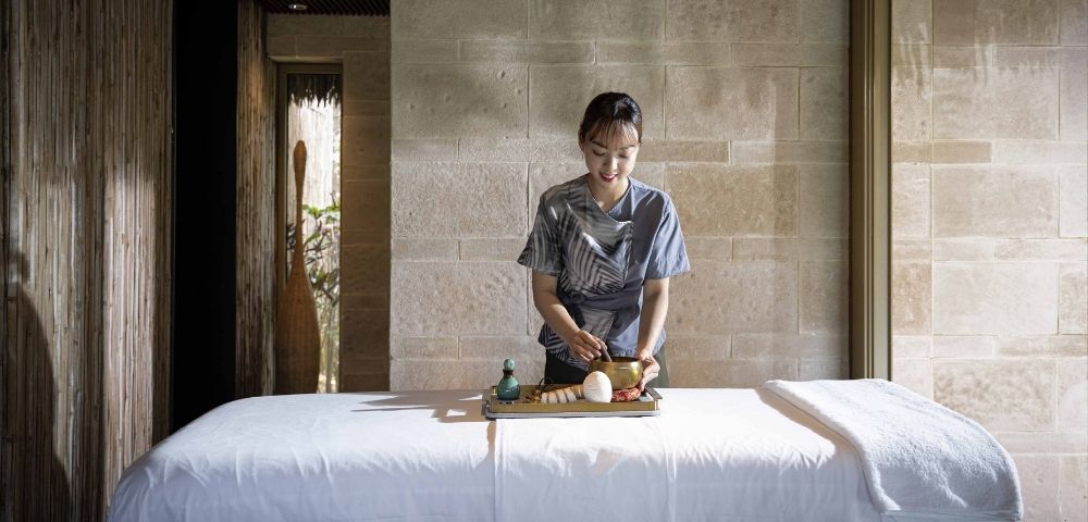 A serene room with a massage table draped in white linens. A person arranges spa items with care, surrounded by soft natural light and wooden decor.