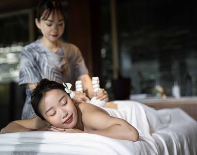 A relaxed woman enjoys a massage in a dimly lit spa, lying on a table with a flower in her hair. The serene therapist uses relaxing tools on her back.
