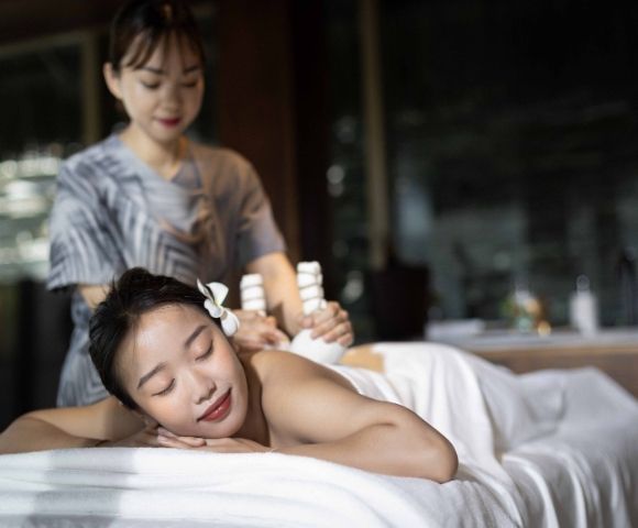 A relaxed woman enjoys a massage in a dimly lit spa, lying on a table with a flower in her hair. The serene therapist uses relaxing tools on her back.