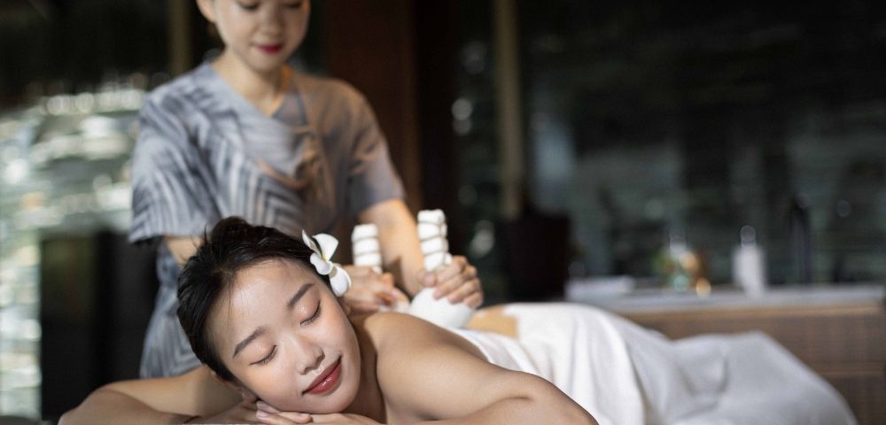 A relaxed woman enjoys a massage in a dimly lit spa, lying on a table with a flower in her hair. The serene therapist uses relaxing tools on her back.
