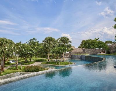 Tropical resort scene with a large, curved pool in the foreground, surrounded by lush greenery and palm trees under a partly cloudy blue sky. Feels serene.