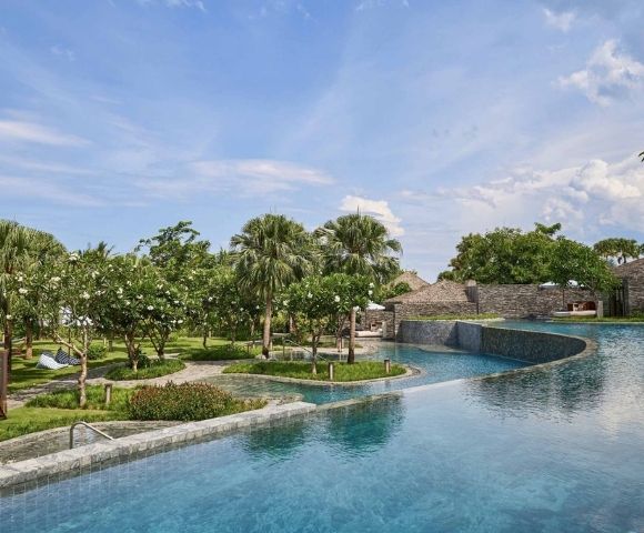 Tropical resort scene with a large, curved pool in the foreground, surrounded by lush greenery and palm trees under a partly cloudy blue sky. Feels serene.