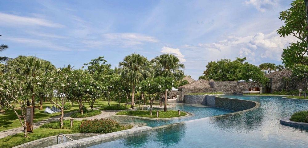 Tropical resort scene with a large, curved pool in the foreground, surrounded by lush greenery and palm trees under a partly cloudy blue sky. Feels serene.