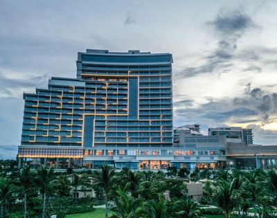 Modern, multi-story hotel at dusk with soft interior lights against a cloudy sky. Foreground features lush greenery and palm trees, evoking a tropical vibe.
