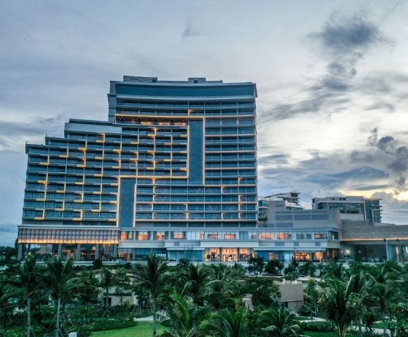 Modern, multi-story hotel at dusk with soft interior lights against a cloudy sky. Foreground features lush greenery and palm trees, evoking a tropical vibe.