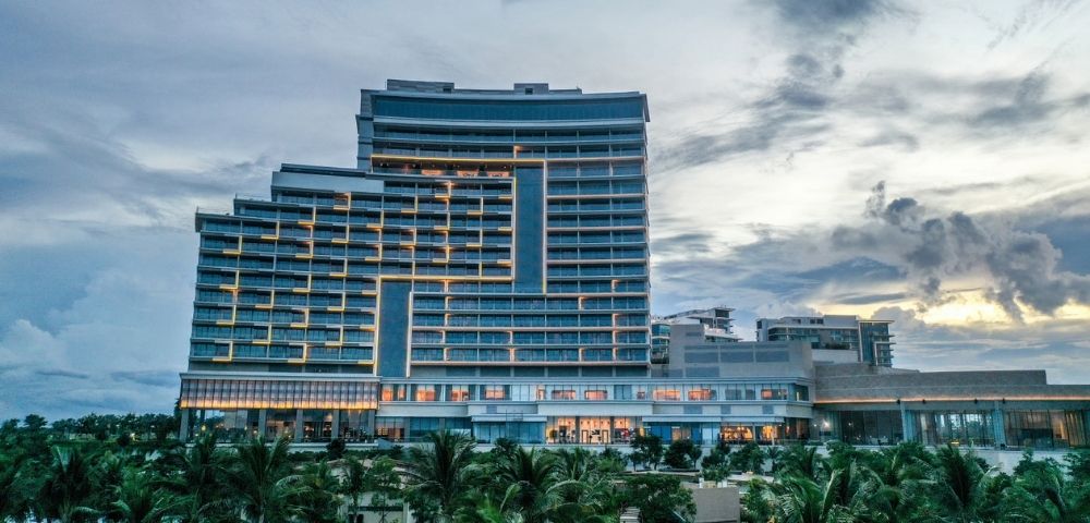 Modern, multi-story hotel at dusk with soft interior lights against a cloudy sky. Foreground features lush greenery and palm trees, evoking a tropical vibe.