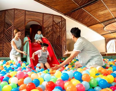 A joyful family plays in a ball pit. A child slides down a red slide into colorful balls, with parents excitedly waiting. The setting is bright and playful.