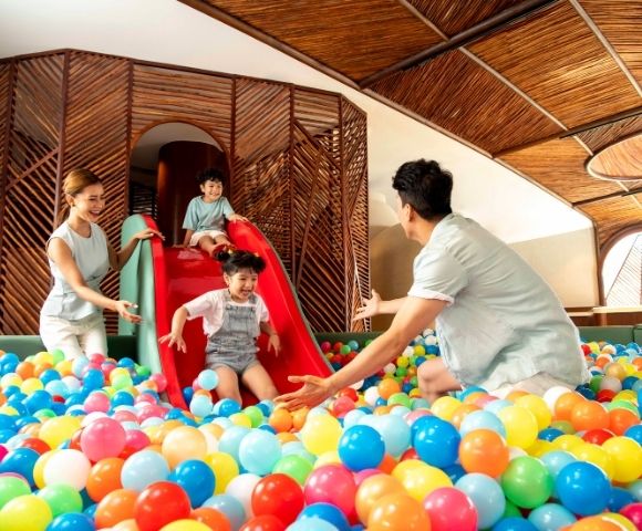 A joyful family plays in a ball pit. A child slides down a red slide into colorful balls, with parents excitedly waiting. The setting is bright and playful.