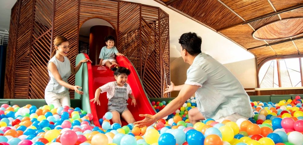 A joyful family plays in a ball pit. A child slides down a red slide into colorful balls, with parents excitedly waiting. The setting is bright and playful.