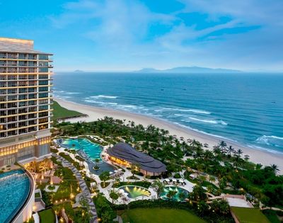 Luxurious beachfront resort with a modern high-rise, lush gardens, and winding pools. Overlooks a serene beach and calm ocean under a clear blue sky.