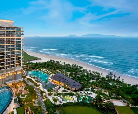 Luxurious beachfront resort with a modern high-rise, lush gardens, and winding pools. Overlooks a serene beach and calm ocean under a clear blue sky.