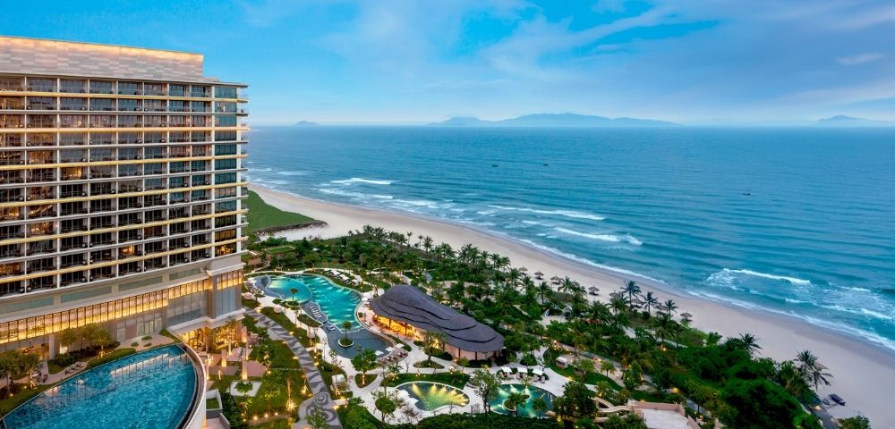 Luxurious beachfront resort with a modern high-rise, lush gardens, and winding pools. Overlooks a serene beach and calm ocean under a clear blue sky.