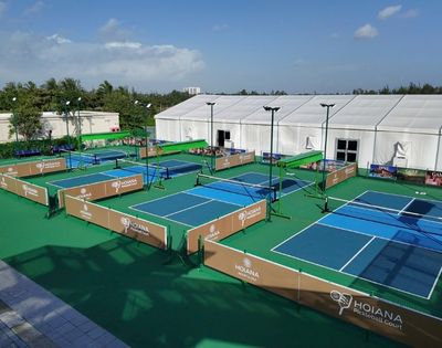 Tennis complex with multiple courts enclosed by brown barriers under a clear blue sky. A large white tent and palm trees are visible in the background.