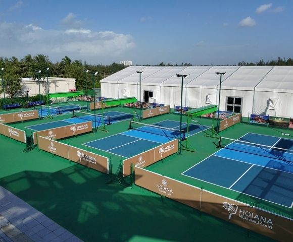 Tennis complex with multiple courts enclosed by brown barriers under a clear blue sky. A large white tent and palm trees are visible in the background.